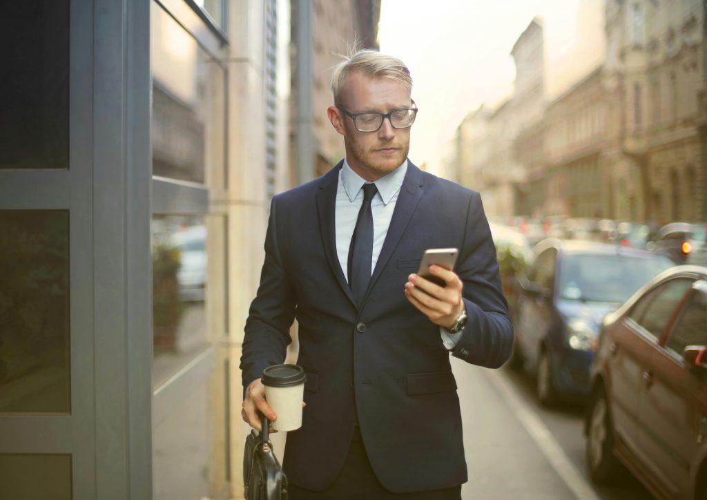 pexels-photo-3771076-3771076 Professional businessman in formal wear walking on a city street while texting on his phone and holding coffee.